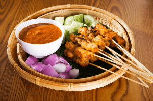Bol contenant une dizaine de brochettes de suya, accompagné de morceaux de concombres et de morceaux d'oignon rouge, de même qu'un petit bol de sauce.